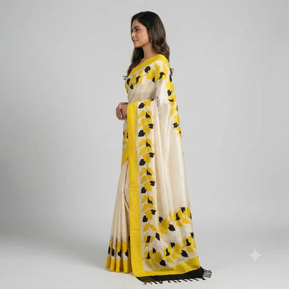 A woman modeling a cream-colored saree with a vibrant yellow border and black floral motifs, photographed against a neutral light gray studio background.