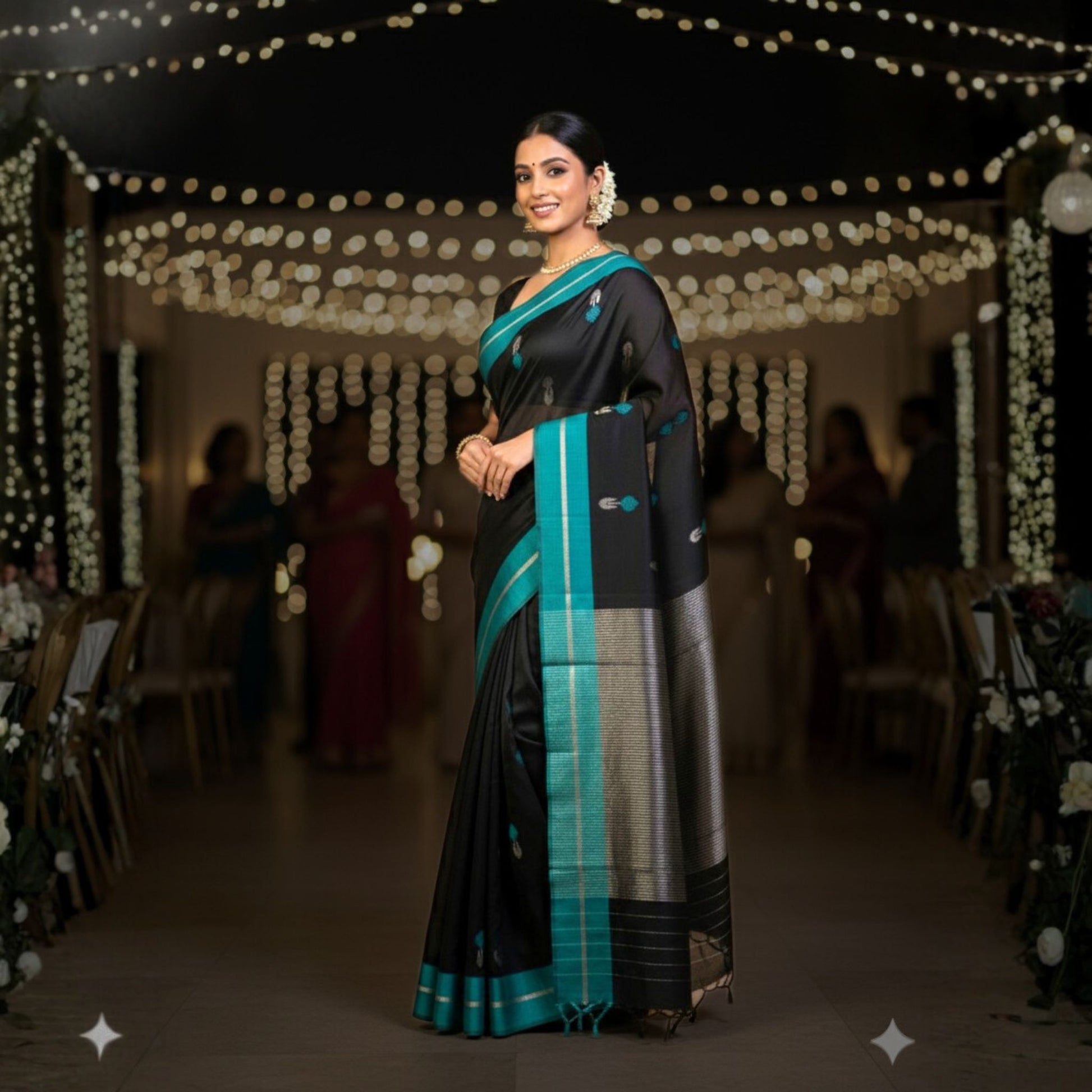 Woman in a black and teal saree standing in a decorated indoor setting with string lights.