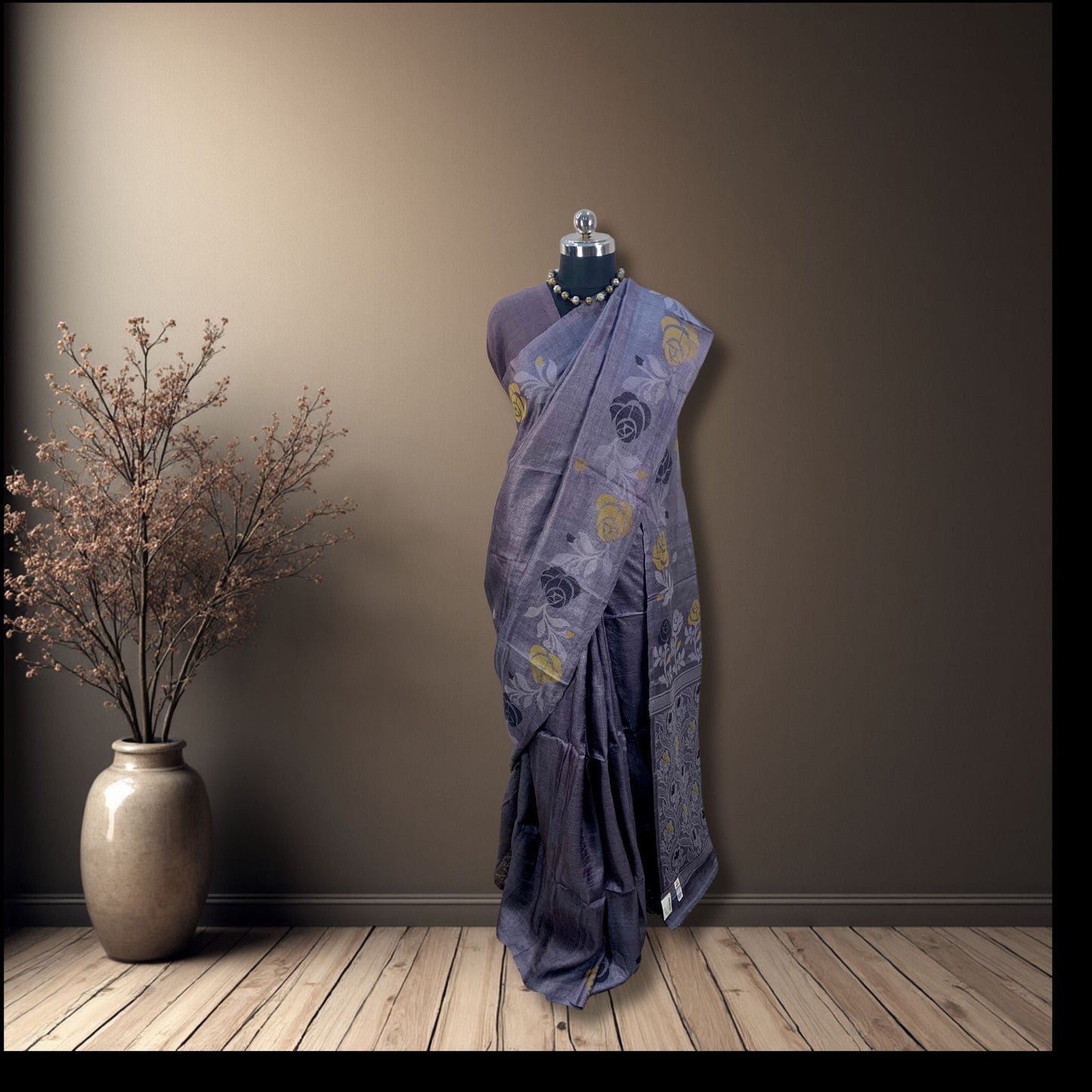 A draped steel grey saree with yellow and black floral motifs displayed on a mannequin torso, set against a dark brown wall and light wood floor, with a decorative vase of dried branches on the left.