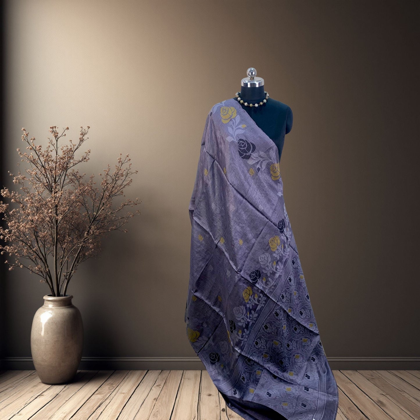 A draped lsteel grey saree with yellow and black  floral motifs displayed on a black mannequin, set against a warm brown wall and wooden floor with a decorative vase on the left.