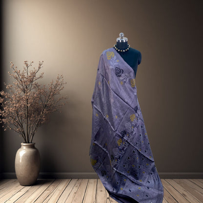 A draped lsteel grey saree with yellow and black  floral motifs displayed on a black mannequin, set against a warm brown wall and wooden floor with a decorative vase on the left.