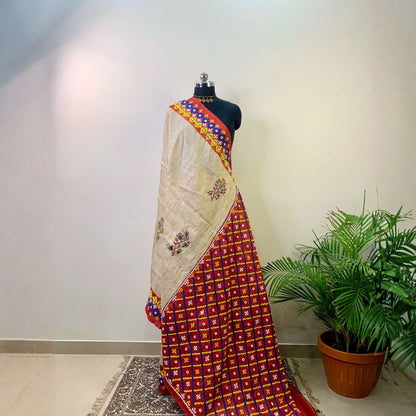 A red and beige patterned saree draped on a black mannequin, set against a plain wall with a potted green plant on the right.