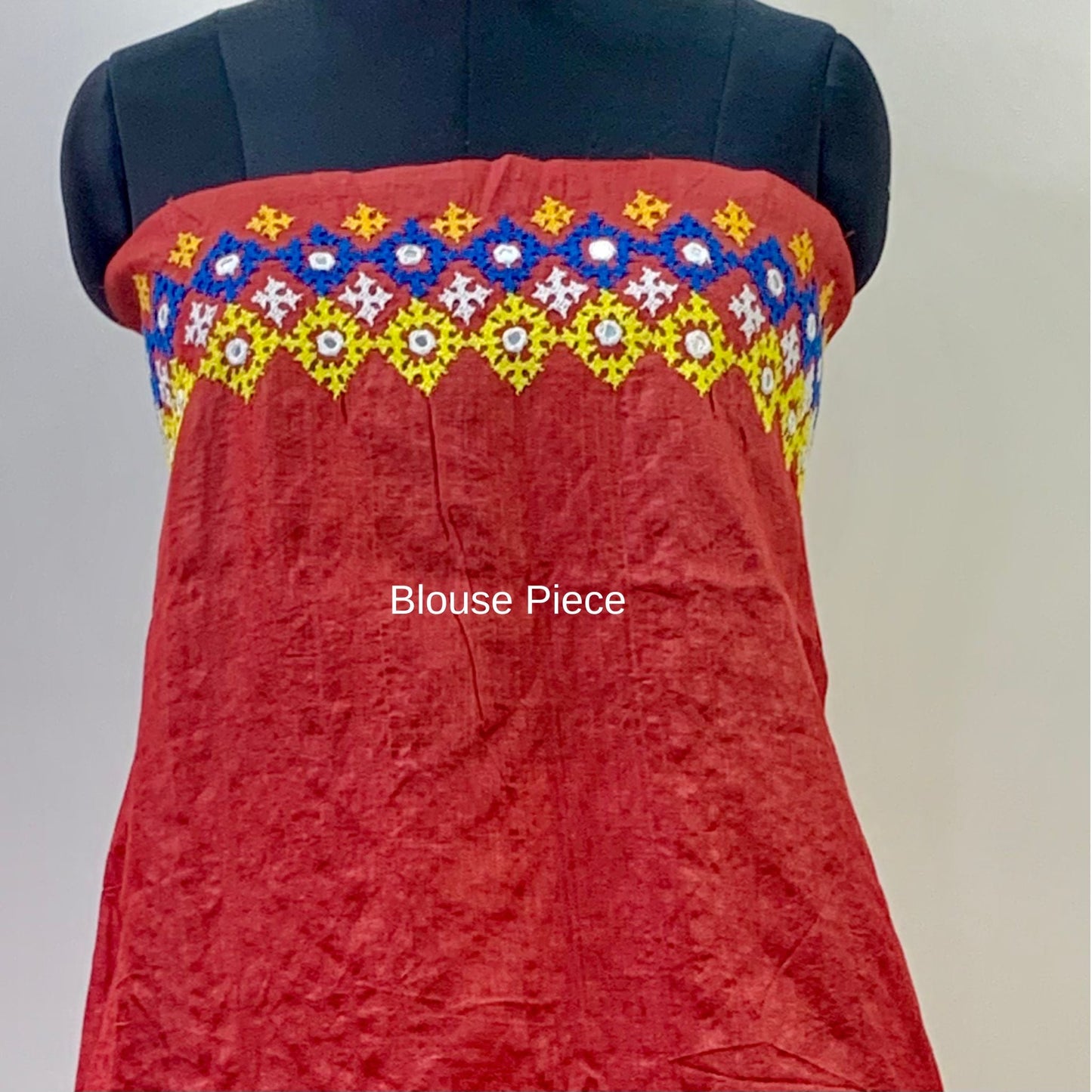 A red cotton blouse piece displayed on a black mannequin torso, featuring a colorful embroidered yoke with geometric patterns in blue, yellow, white, and red.