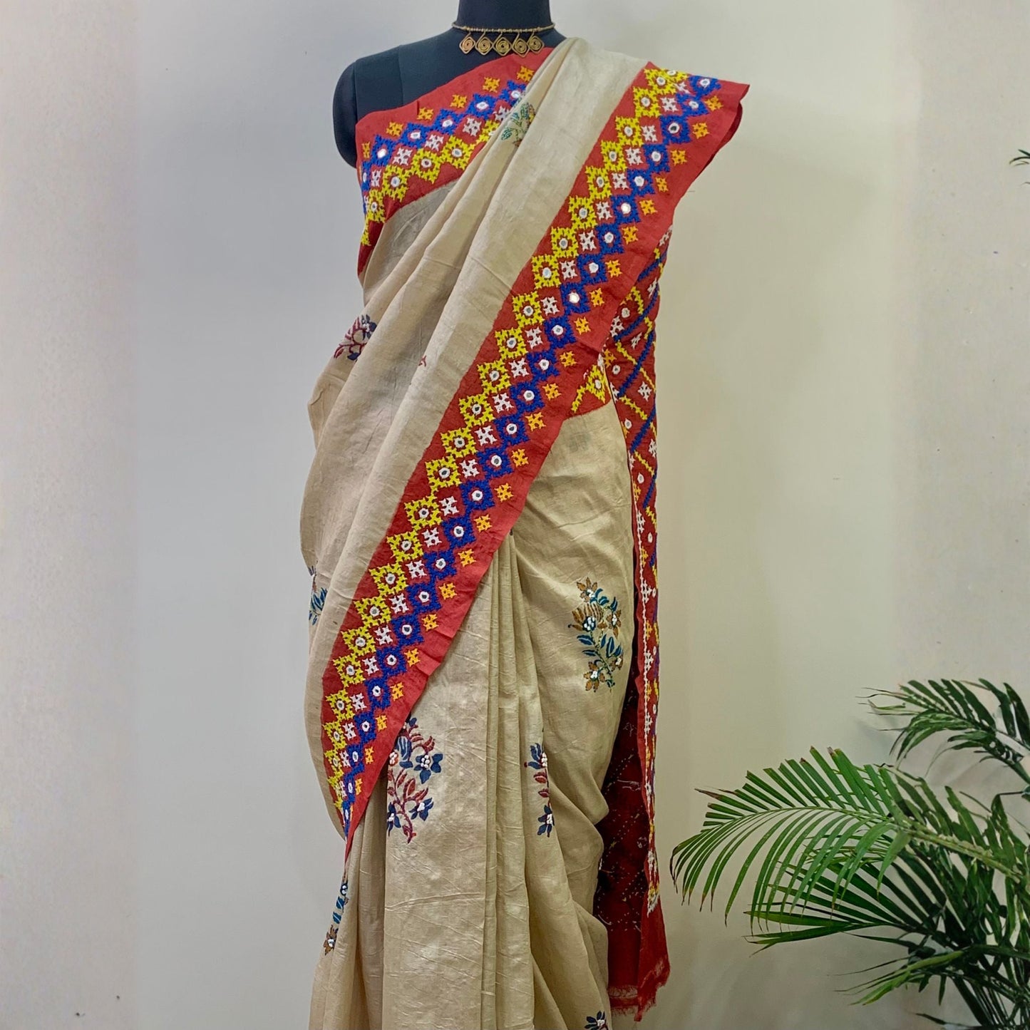 A beige silk saree with a red border decorated with colorful floral patterns, displayed on a black mannequin torso against a plain wall with a green plant in the corner.